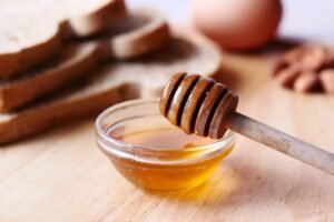 Close-up of honey dipper in a bowl with honey on wooden table beside sliced bread.