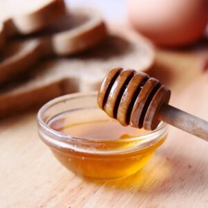 Close-up of honey dipper in a bowl with honey on wooden table beside sliced bread.