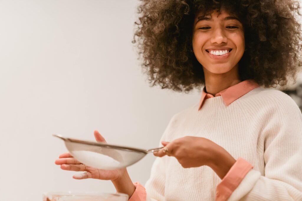 Happy woman with an afro sifting flour in a cozy kitchen setting, enjoying baking.
